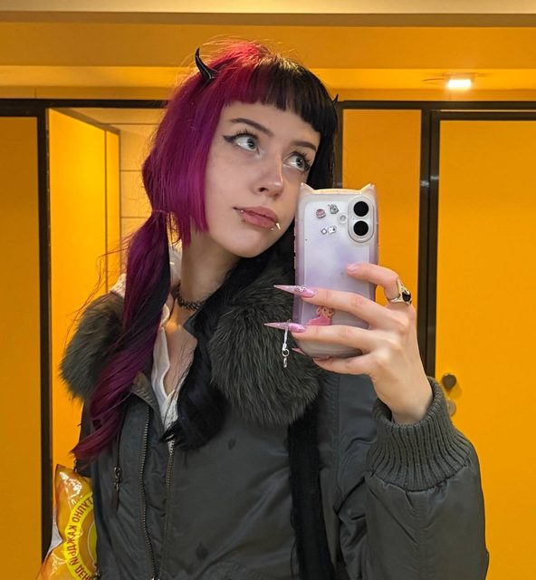 A mirror selfie of a person with magenta and black hair and devil-horn clips, holding a pink cat-ear phone case against their cheek in front of yellow doors.