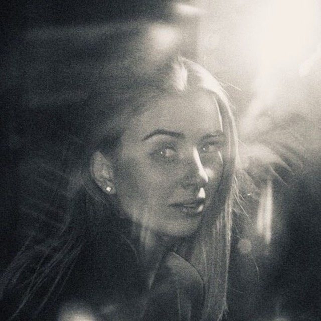 A moody black-and-white close-up portrait of a woman with long hair, lit by a bright flare of light at the upper right, with heavy grain and a soft, hazy look.