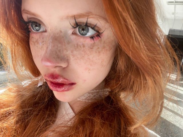 An extreme close-up selfie of a red-haired person with freckles, dramatic lashes and glossy red lips, in bright light.