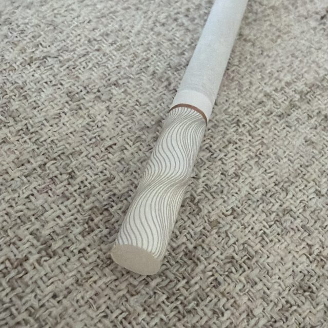 A close-up photo of a white cigarette with a decorative wavy-patterned filter lying on a beige woven fabric surface.