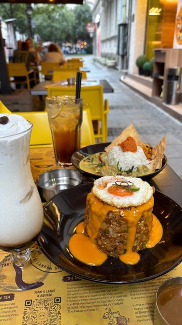A photo of a meal at an outdoor café table: a tall creamy iced coffee drink, an iced tea with lemon, a plate of fried rice cake topped with a fried egg and cheese sauce, and a bowl of curry with rice and papadum.