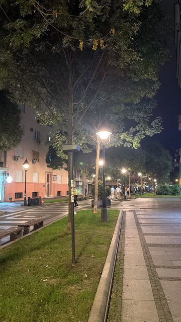 A night photo of a quiet pedestrian street with tall trees, glowing lamp posts, benches, a lawn strip and a pink apartment building. A few people walk in the distance.