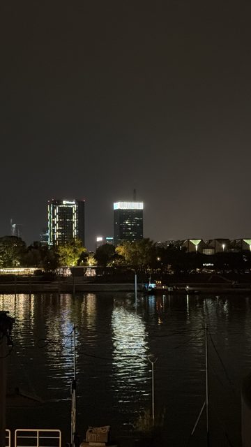 A night photo across a river or harbor toward a lit city skyline, with two illuminated high-rise towers, trees and low buildings on the far bank, and light reflections on the dark water.