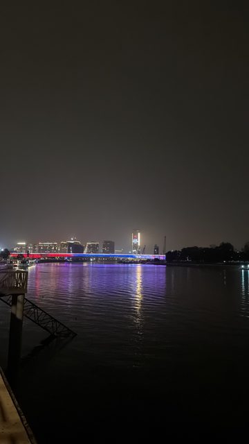 A night photo of a wide river with a long bridge lit in red, blue and white and a city skyline with a lit tower on the far bank.