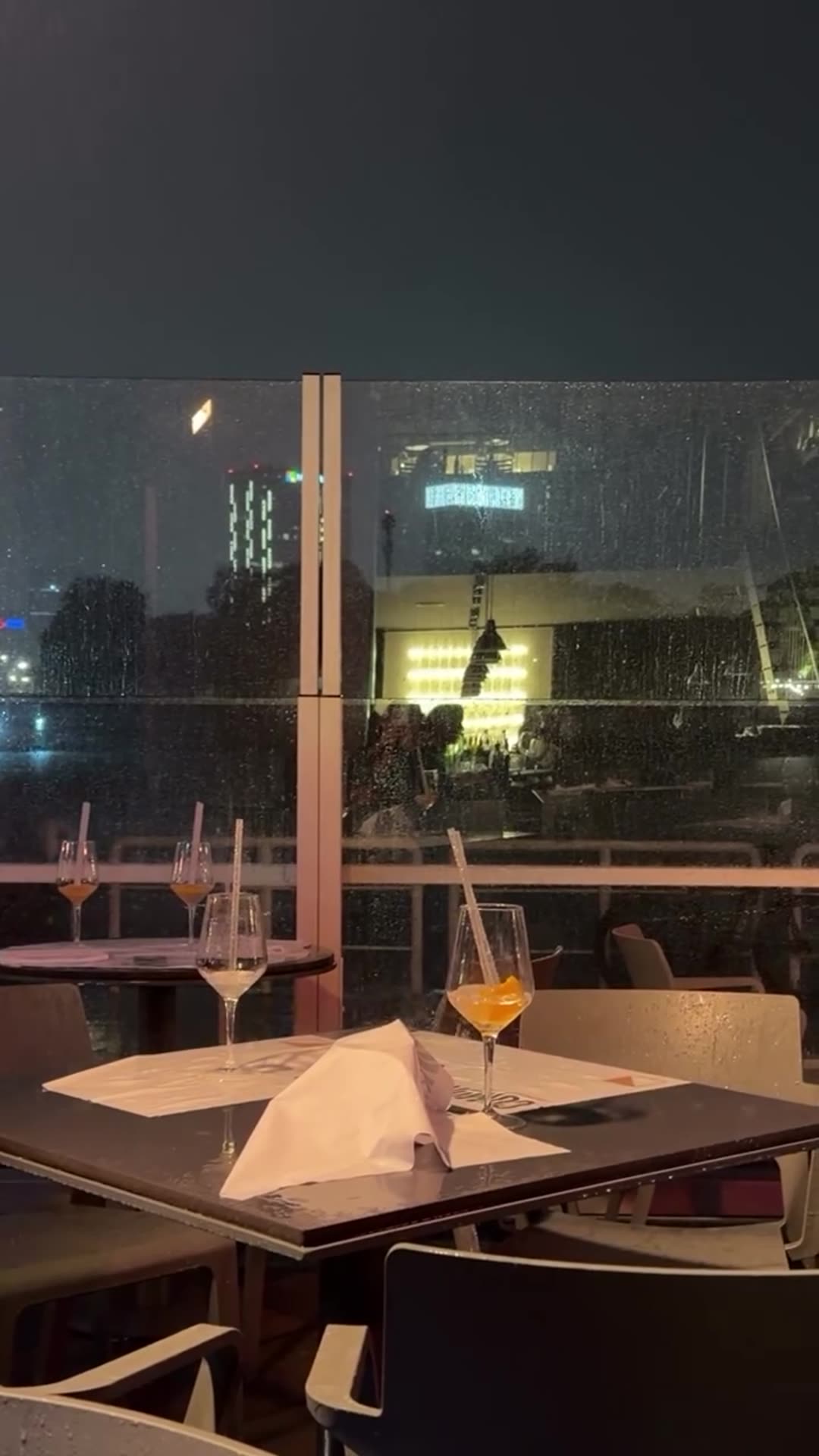 A short, silent story video shows a restaurant terrace table at night behind a rain-spotted glass wall, with two orange spritz-style drinks and wine glasses with straws, and a folded napkin. A lit bar and city lights are visible beyond the glass.