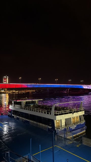 A night photo of a moored river boat named Horizont (Beograd) on a wet dock, with a bridge lit in red, blue and white across the river behind it.