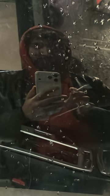 A dark, blurry selfie taken through a rain-covered reflective surface, showing a hooded person with a phone and a cigarette.