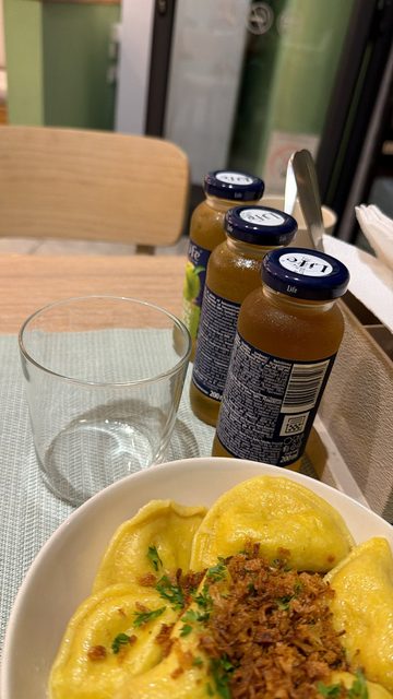 A story photo of a plate of yellow filled dumplings (probably pierogi or ravioli-style) topped with fried onion and parsley, next to three small bottles of Life juice and an empty glass on a restaurant table.