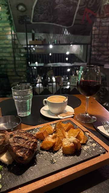 A story photo of a restaurant table at night with a slate board of grilled steak medallions and roasted potato wedges sprinkled with grated cheese, next to a glass of red wine, a cup of coffee and a glass of water.
