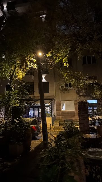 A night photo of a street lamp glowing among autumn trees on a quiet street, with a lit café terrace and a parked car in the background.