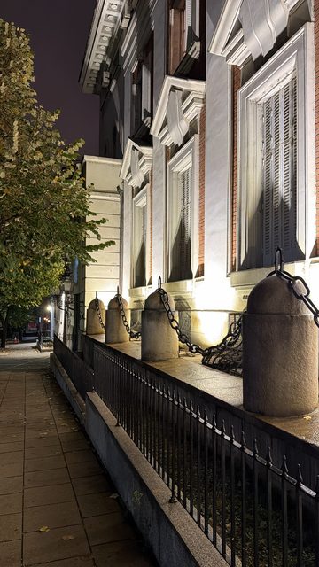 A night photo of a floodlit neoclassical building with shuttered windows, stone bollards linked by chains and an iron fence, beside a paved sidewalk and a tree with yellowing leaves.
