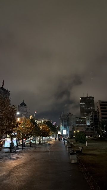 A story photo of a wide pedestrian boulevard at night under a heavy dark cloudy sky, with autumn trees, benches, street lamps and illuminated historic and modern buildings along it.