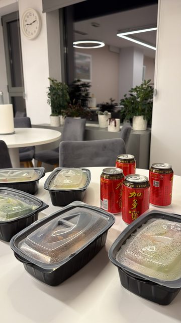 A story photo of five black takeaway food containers with condensation-fogged lids and four red cans of Jia Duo Bao herbal tea on a white round table in a modern office or common room.
