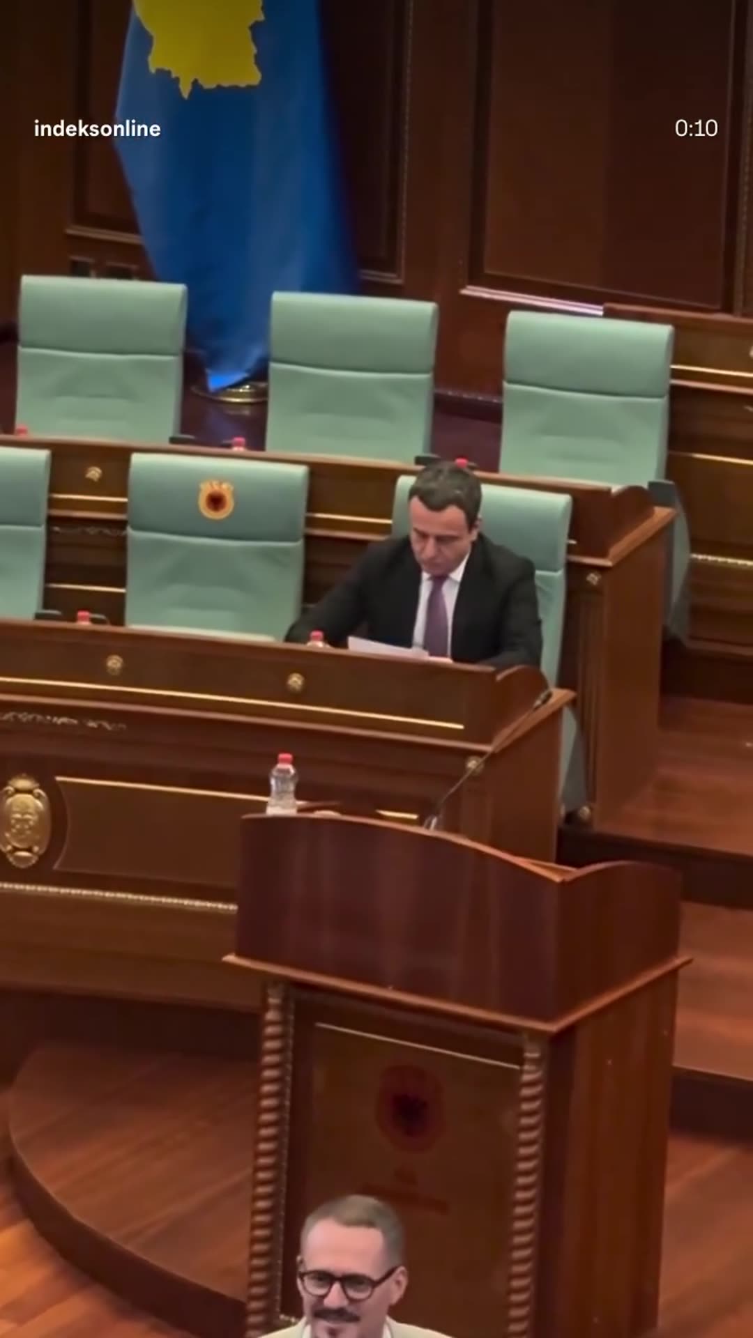 A story reshares an IndeksOnline reel showing Albin Kurti arriving at his seat in the Kosovo Assembly chamber, sitting down and reading documents. The camera finally pulls back to a wide view of the half-empty hall with deputies milling around.