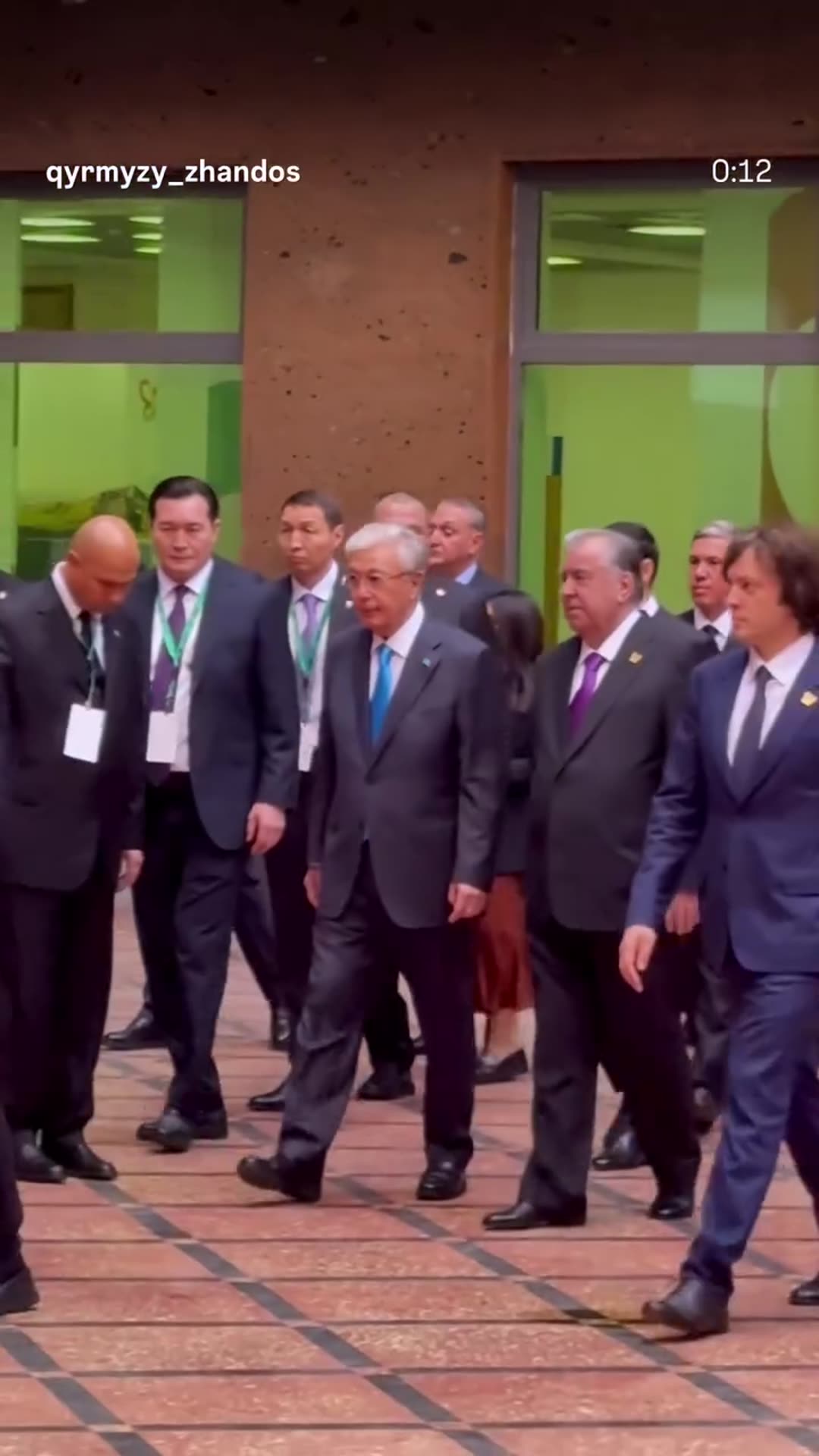 A story reshares a vertical reel filmed at a summit venue. A group of suited officials walks across a brick-paved courtyard past brown stone walls and green-glass windows, while staff and journalists look on.