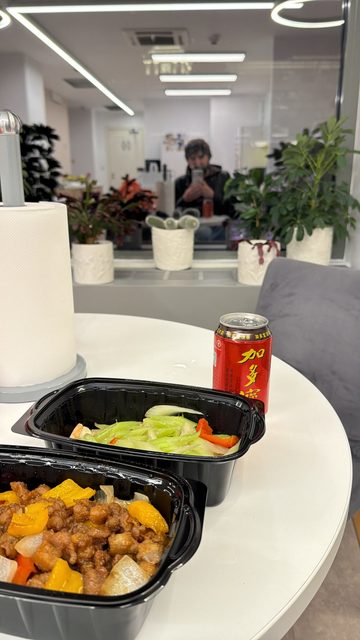 A story showing two takeaway containers of Chinese food, one with sweet-and-sour style crispy pork and peppers and one with stir-fried celery and red pepper, next to a red can of Jiaduobao herbal tea on a white round table, with a mirror selfie in the background.