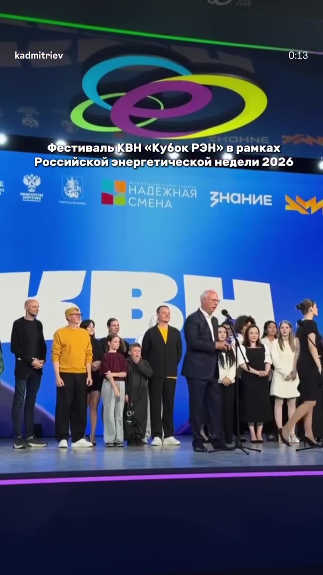 A reshared reel from @kadmitriev showing highlights of the KVN 'Cup of RAEN' festival held during Russian Energy Week 2026. It shows a selfie with a participant, the audience, panel members seated at the front, a speech on stage, an embrace on stage with a young man, and a wide shot of the hall.