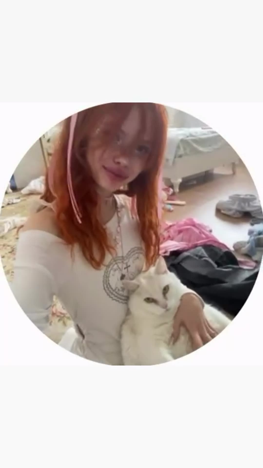 A circular-cropped video clip of a young woman with red hair in a messy bedroom, holding a white cat, smiling at the camera and taking a mirror selfie with her phone.