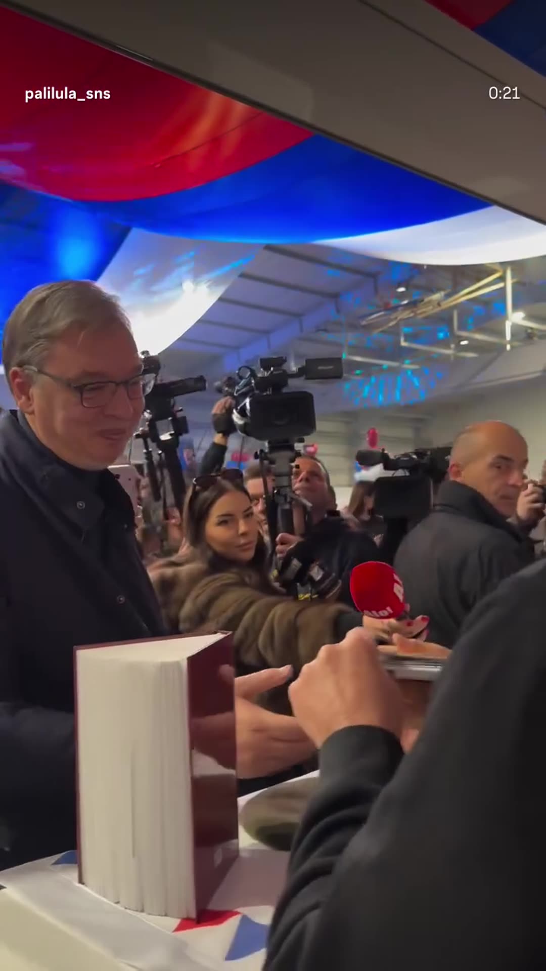A story reshares a reel from @palilula_sns showing a man in a dark jacket touring a busy fair with stalls. He stops at a stall with bottles of spirit and traditional hats, then at a book stand where he looks through and signs a book while TV crews film him.