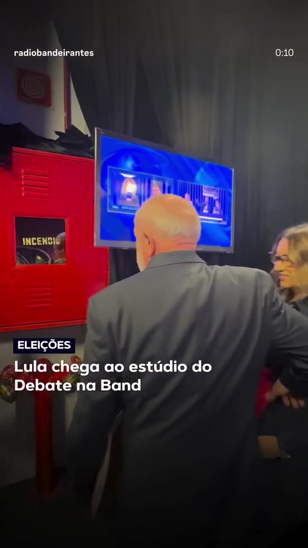 A reshared Rádio Bandeirantes reel shows Lula and his entourage walking backstage to the Band studio for the 2026 election debate. A reporter narrates over the footage.