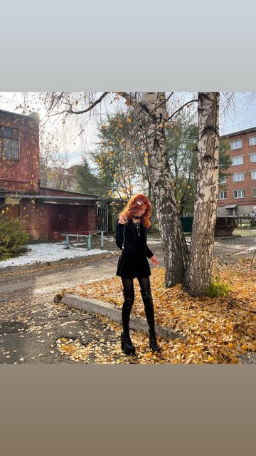 A story photo of a young woman in an all-black outfit standing under birch trees among yellow autumn leaves, between red-brick buildings, with patches of early snow on the ground.