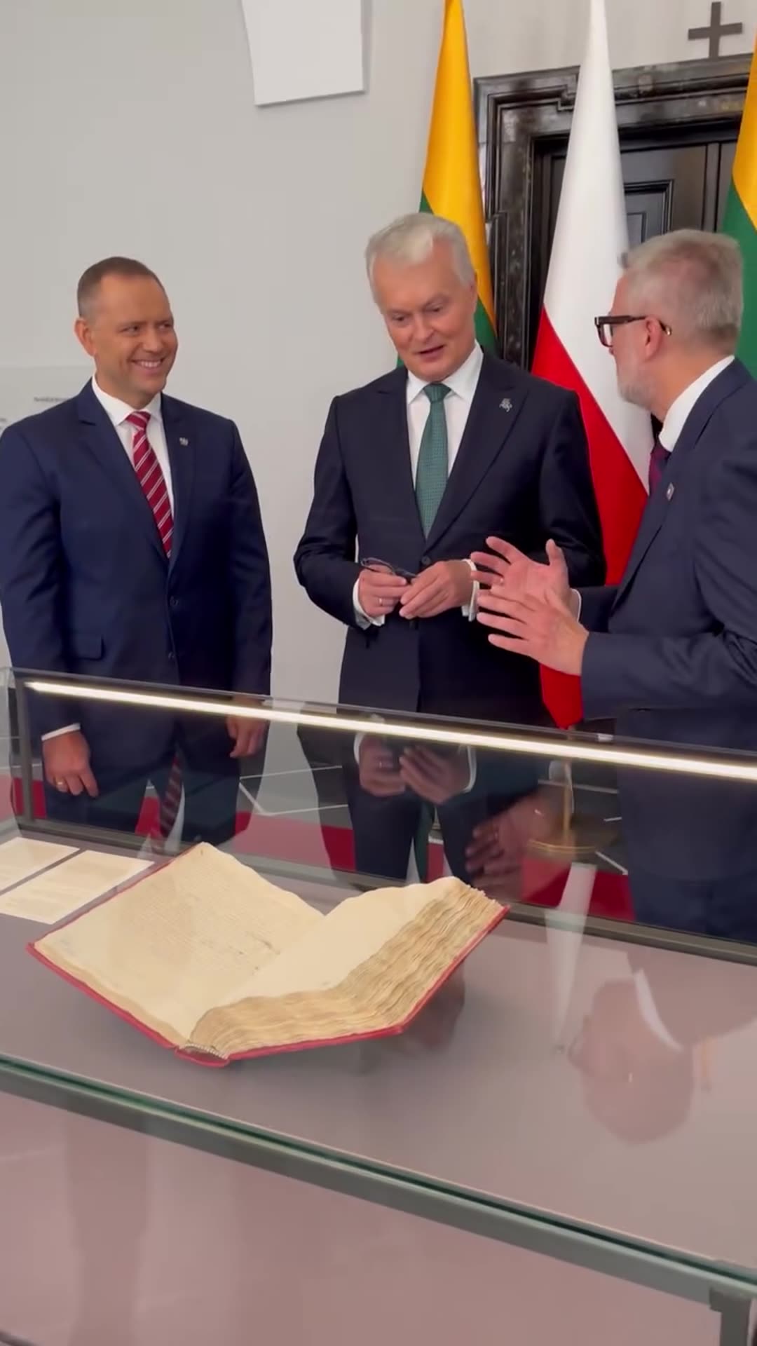 A reshared story shows an old open manuscript book in a glass display case, then three men in suits chatting beside it, with Lithuanian and Polish flags behind them.