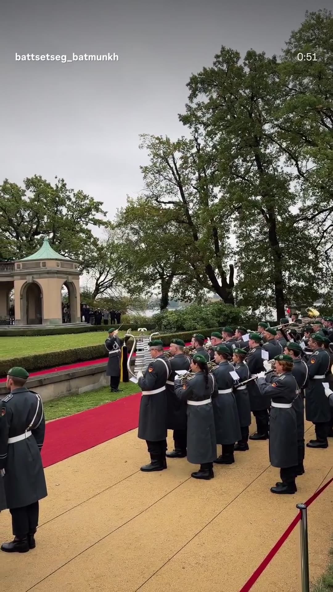 A story reshares a reel of the military welcome ceremony on a red carpet at a villa, with a band playing, the German flag raised alongside another flag, and two leaders standing at attention.