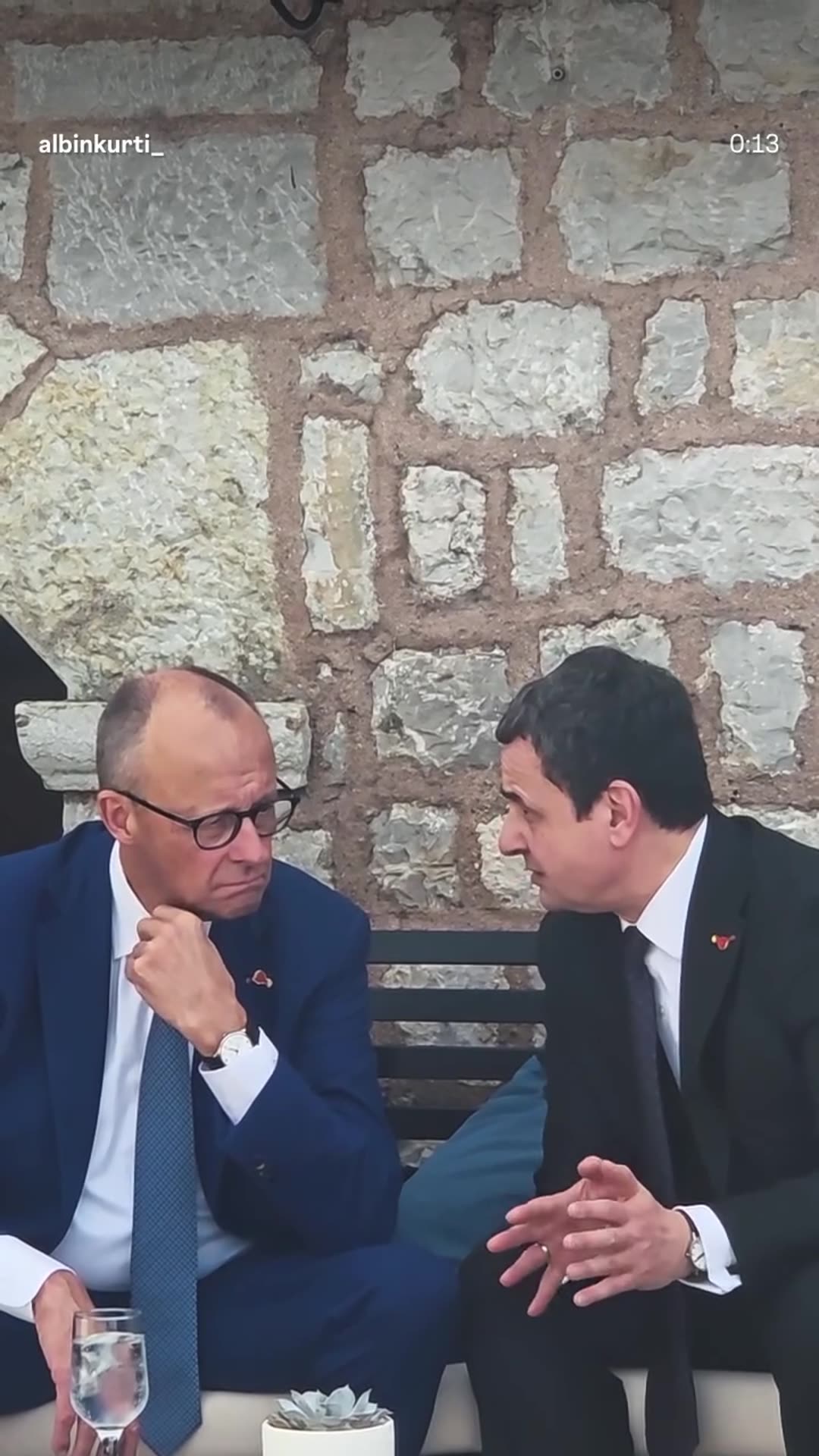 A story reshares Albin Kurti's reel from the 13th Berlin Process summit. It shows his greeting and talks with German Chancellor Friedrich Merz at a stone-walled seaside venue, followed by scenic views of the coast.