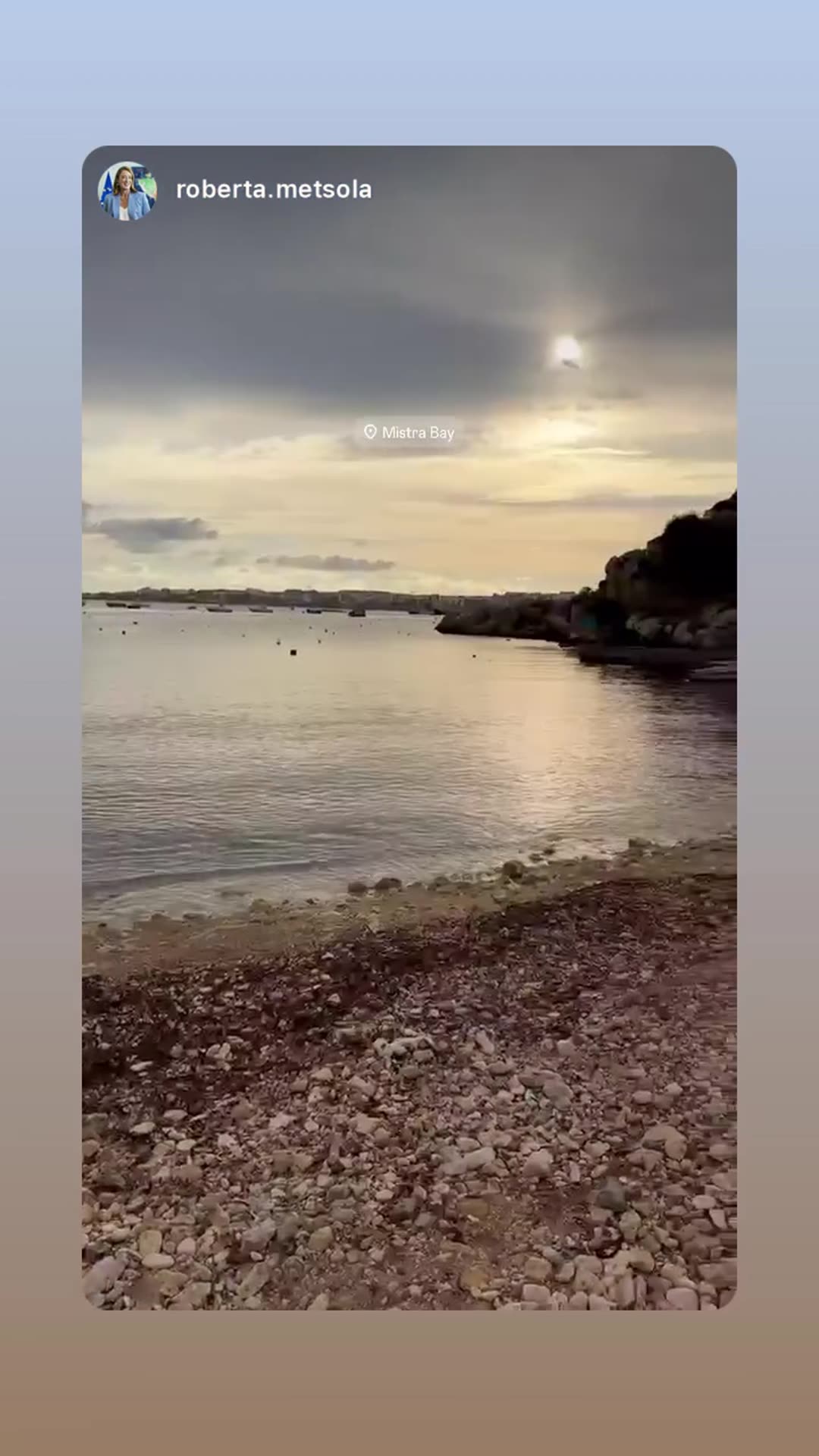 A story reshares a short video by Roberta Metsola, filmed from a pebbly shore at Mistra Bay. It pans across calm water, boats and hills under a cloudy sky, with the sun breaking through at the end.