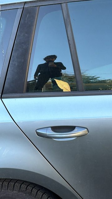 A story photo of the rear door of a light blue-grey car. The window reflects the photographer holding a phone against a blue sky and trees, and a yellow autumn leaf sits on the glass in front of the reflection.