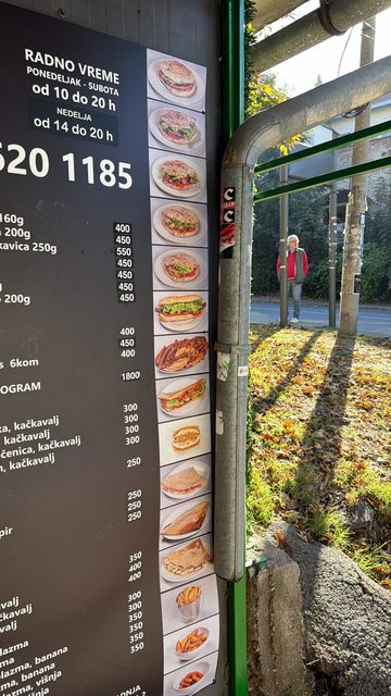 A photo of a Serbian-language fast-food menu board with photos of sandwiches, crepes and fries, with opening hours and prices, next to a green metal frame and autumn leaves.