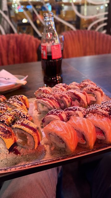 A close-up photo of a platter of sushi rolls (salmon nigiri-style rolls, rolls topped with eel sauce and sesame, and torched rolls) next to a glass bottle of Coca-Cola on a wooden table in a restaurant.