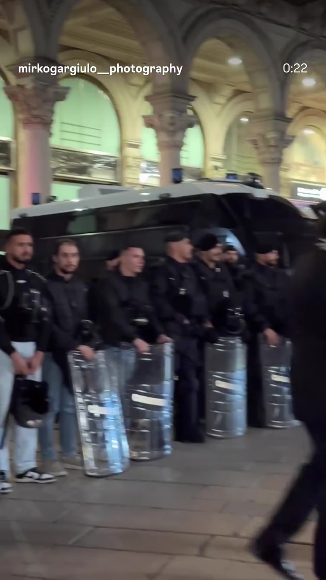 A story reshares a reel filmed at night in Piazza del Duomo, Milan, showing a Carabinieri riot-shield cordon in front of police vans, with a group of people standing beside it and two officers walking past.