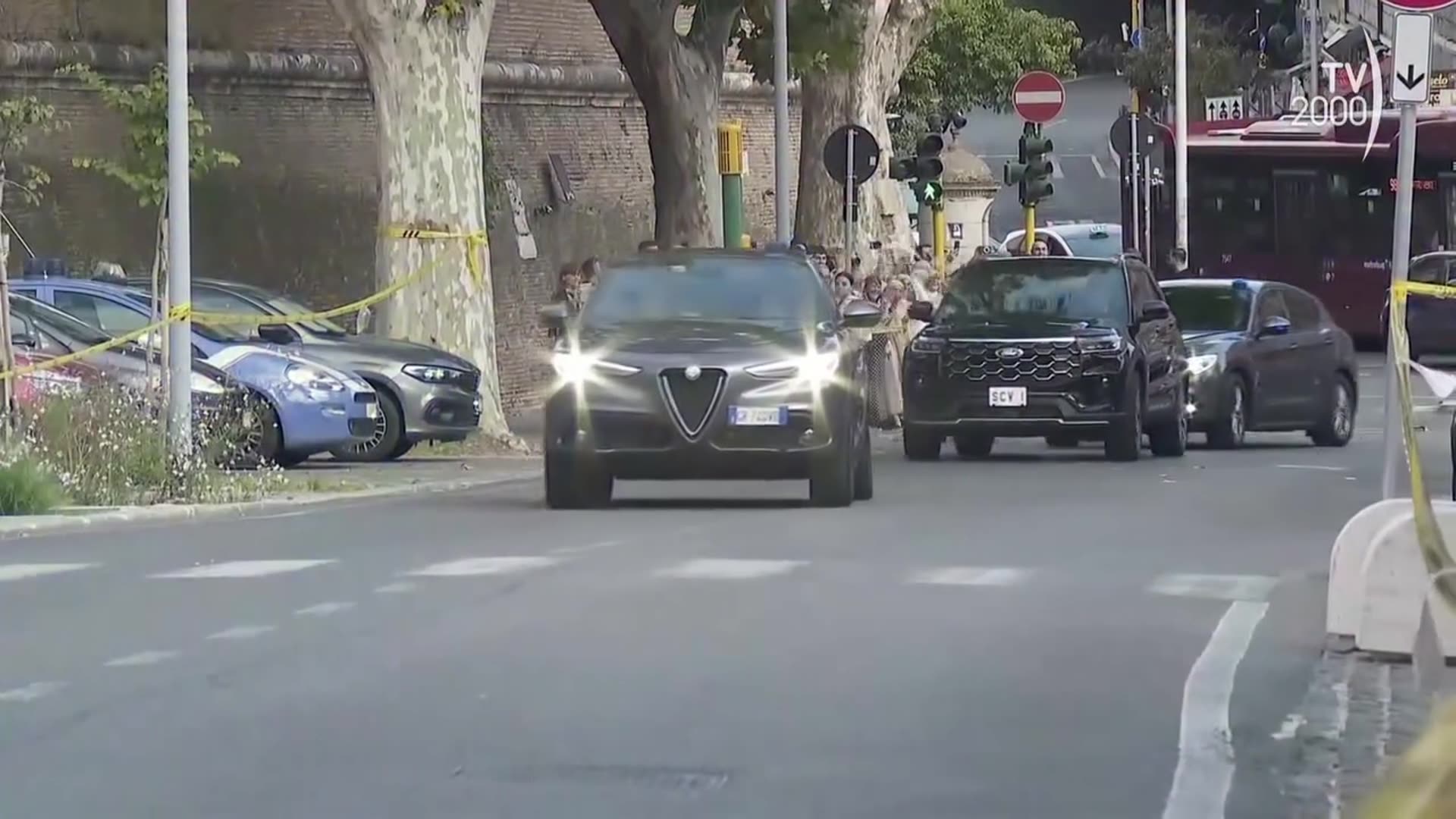 A reshared TV2000 news clip shows a motorcade with police escort cars and a black SUV with Vatican plates (SCV 1) driving through Rome and entering Vatican City, while onlookers and photographers watch. The caption says this is Pope Leo XIV returning home from Gemelli Hospital.