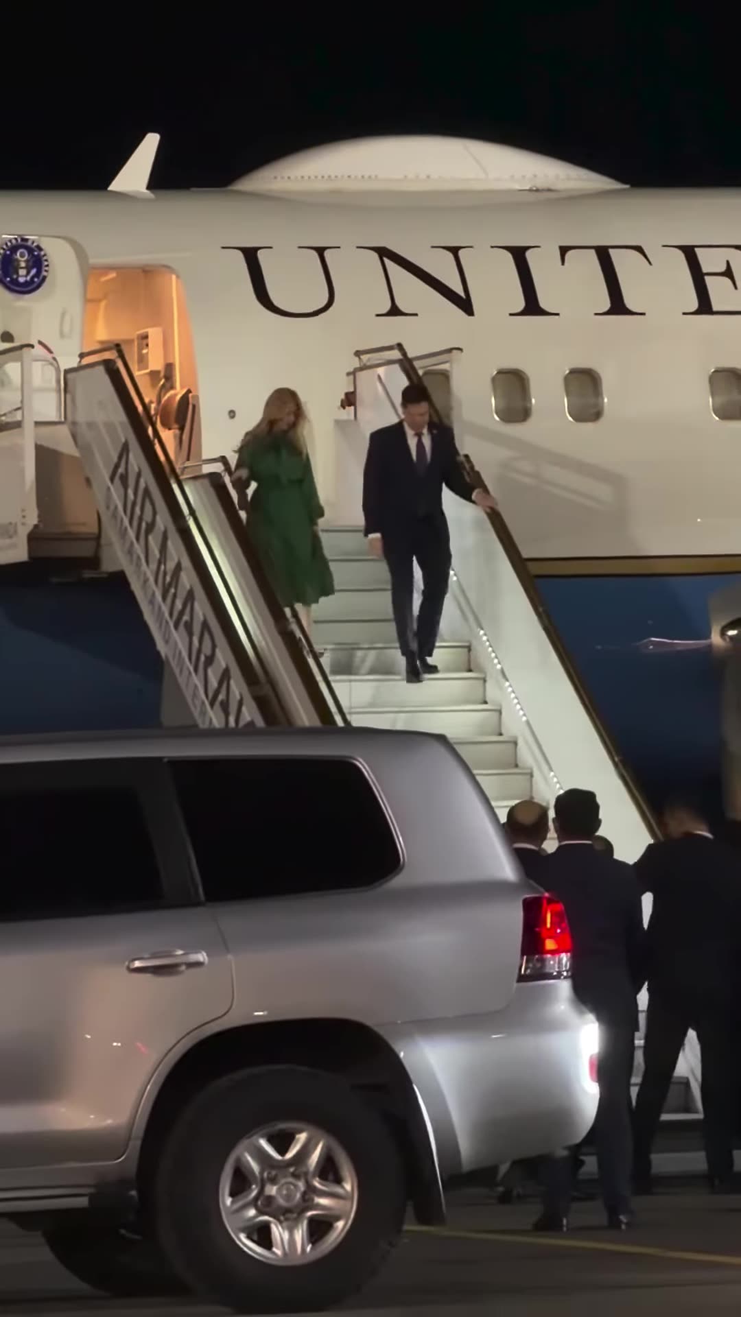 A night-time vertical video, filmed through a car windshield, of passengers disembarking from a US government aircraft marked 'United States of America' at an airport. A group of men in dark suits waits at the foot of the stairs, with SUVs parked nearby. The caption says this is a visit for the Ame…