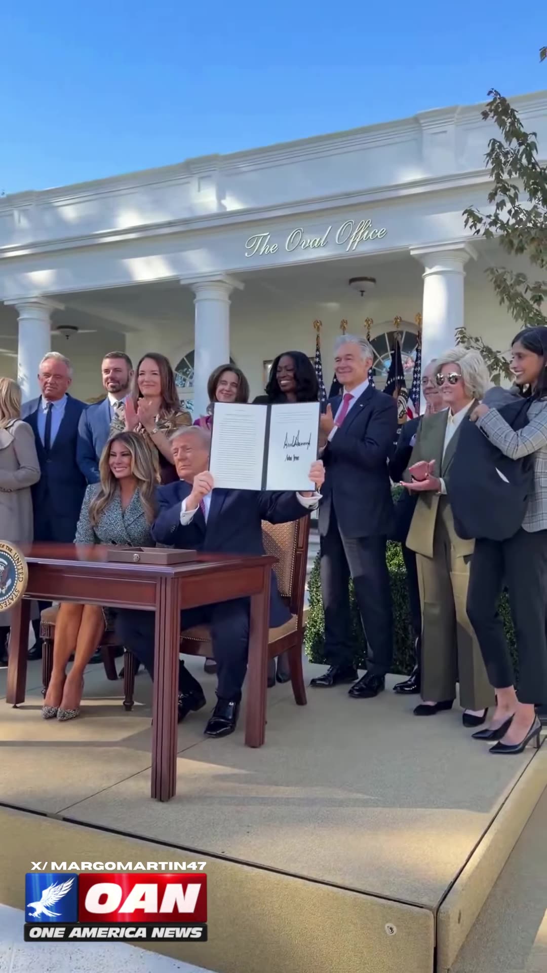 A reshared OAN reel shows President Trump holding up a signed Executive Order with First Lady Melania Trump beside him, while onlookers applaud. The order establishes a Women's Health Division at NIH and a White House Women's Health Task Force.