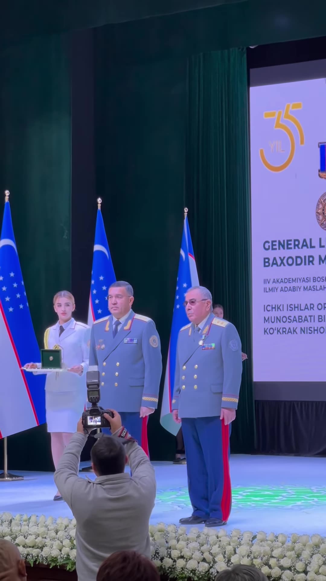 A repost of a reel showing a ceremony on stage where Lieutenant General Bahodir Matlyubov is awarded the "Interior Ministry 35th Anniversary" breast badge. A senior officer pins the badge on him, they pose, and the presenter applauds.